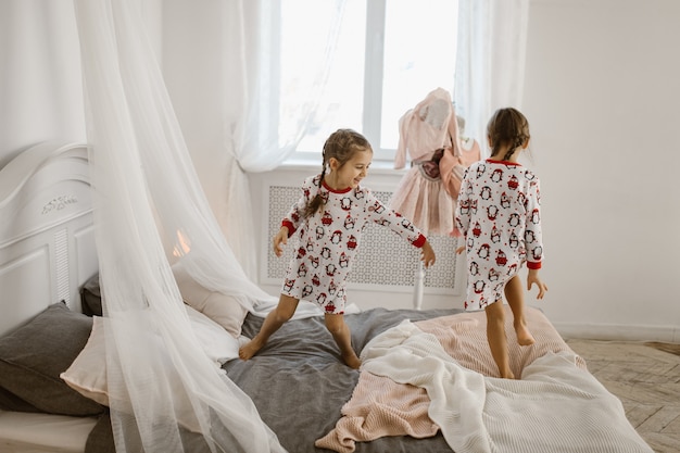 Две маленькие девочки в пижамах весело прыгают на кровати в залитой солнцем уютной спальне. .