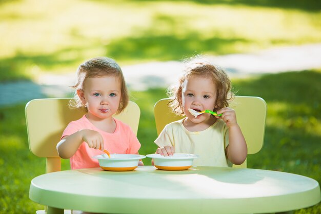 Two little girls sitting at a table and eating together against green lawn