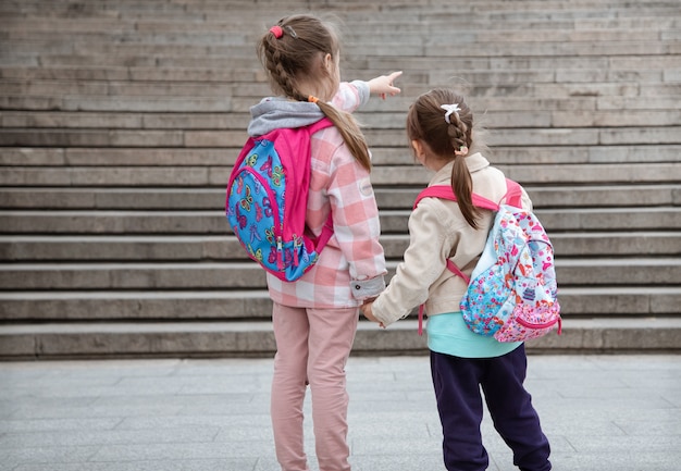Two little girls with backpacks on their backs go to school together hand in hand. Childhood friendship