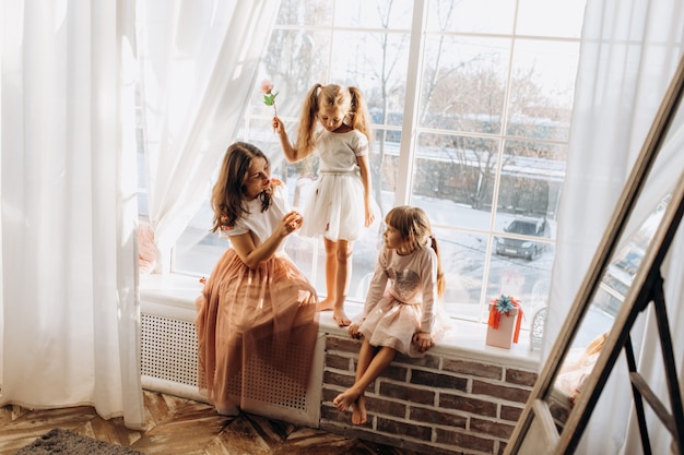 Две сестренки в красивых платьях и молодая мама сидят на подоконнике у зеркала, а на улице зима. Premium Фотографии