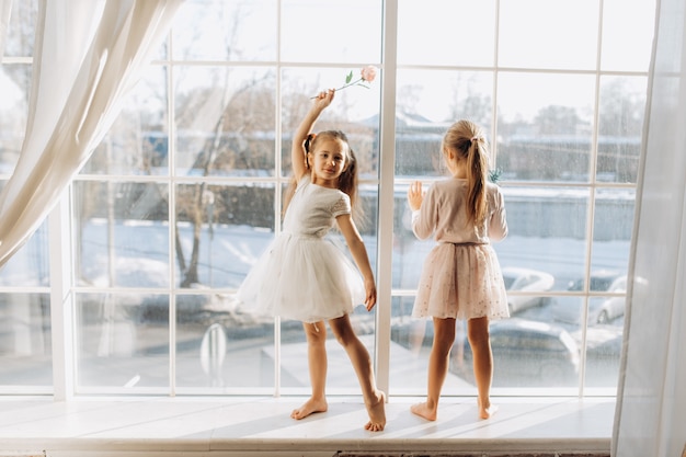 Две сестренки в красивых платьях стоят на подоконнике рядом с зеркалом.