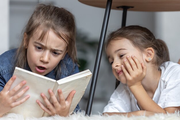 Two little sisters have fun reading a book together while lying on the floor in their room.