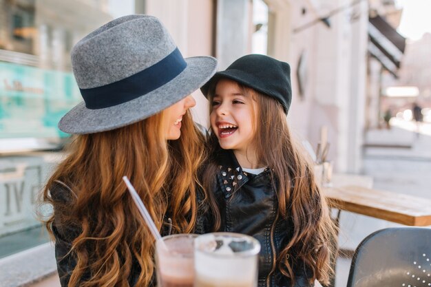 Two long-haired curly sisters looking with love at each other, enjoying sunny morning in outdoor cafe.