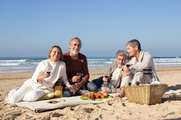 砂浜でピクニックを楽しんでいる2人の素敵な年配のカップル。昔の友達が一緒にぶらぶらして、ワインを飲み、楽しい時間を思い出します。笑顔でカメラを見ている人。レジャー、友情の概念