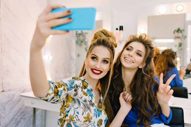 Две милые улыбающиеся молодые женщины веселятся, делая селфи по телефону в парикмахерской
