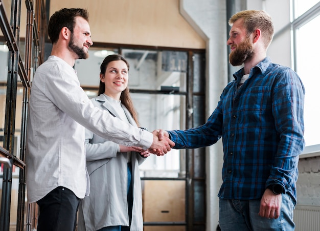 Two male coworker shaking hand in front of smiling businesswoman in office