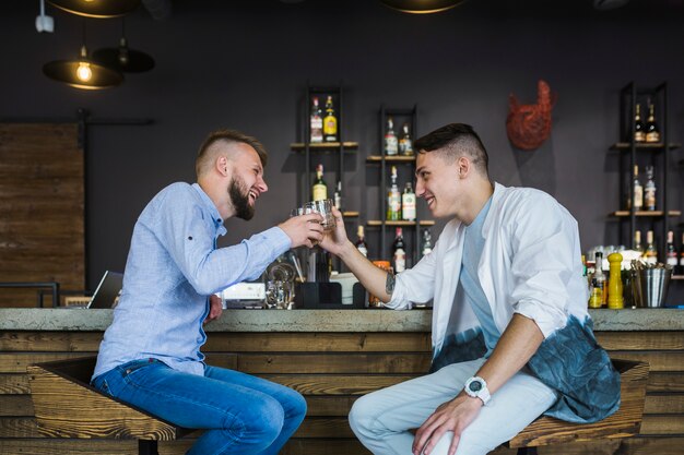 Two male friends sitting at bar counter toasting glasses of drinks