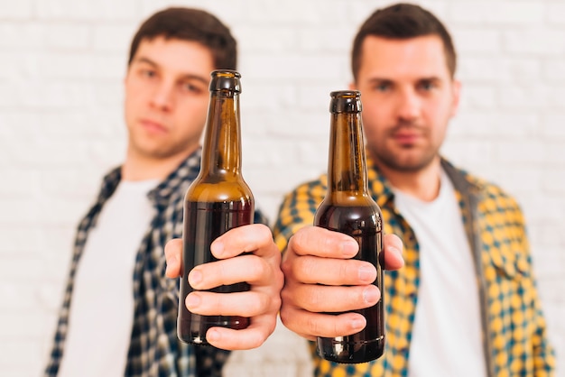 Two male friends standing against white brick wall showing beer bottles toward camera