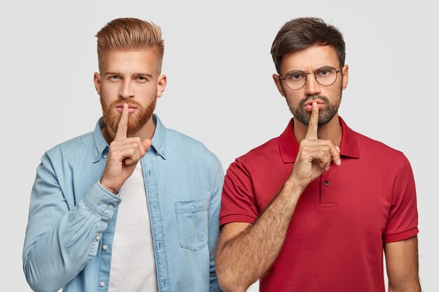 Two male friends with thick beards, keep fore fingers on lips, look secretly, tell very private information, stands next to each other, isolated over white wall. People, secret concept