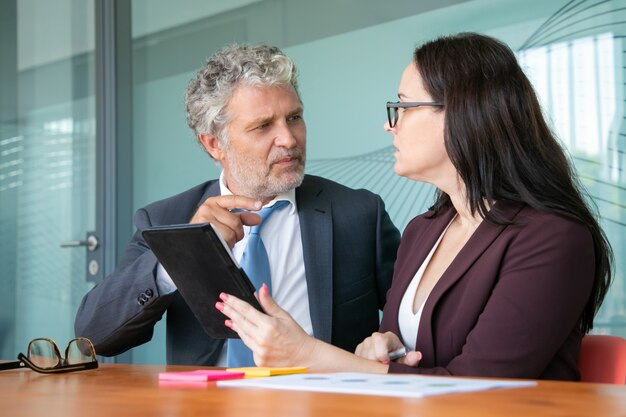 Two managers sitting and talking in meeting room, using tablet, discussing and analyzing reports, arguing