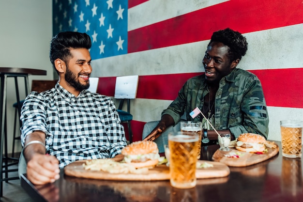 Two men are sitting together in a bar or restaurant lounge. They are laughing and talking while enjoying burgers and beer