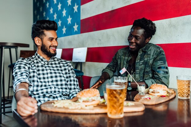 Two men are sitting together in a bar or restaurant lounge. They are laughing and talking while enjoying burgers and beer