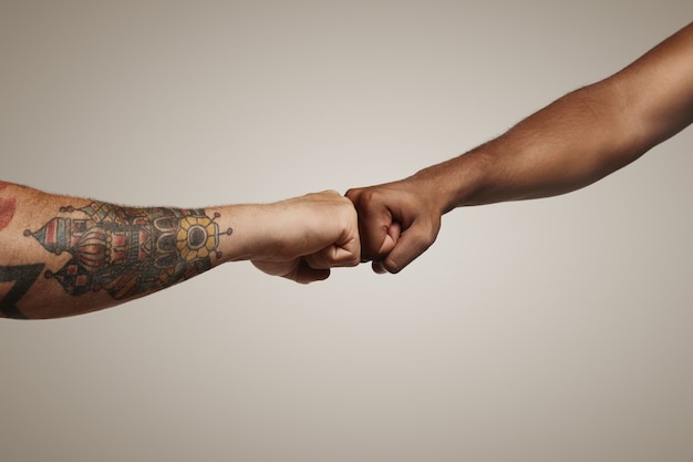 Two men one light skinned with tattoos and another dark skinned do a fist bump on white wall close up