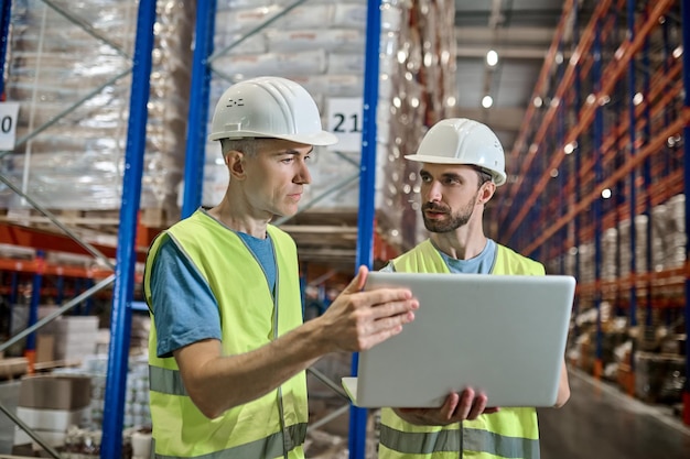 Two men in overalls with laptop talking in warehouse