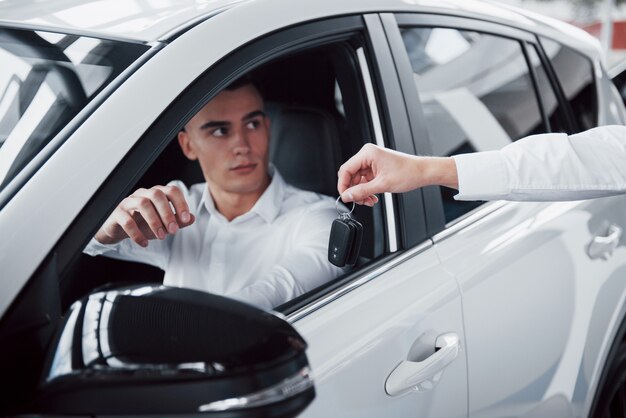 Two men stand in the showroom against cars. Close-up of a sales manager in a suit that sells a car to a customer. The seller gives the key to the customer.