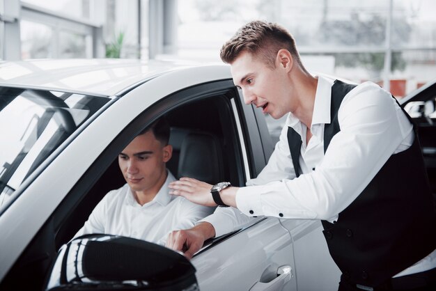 Two men stand in the showroom against cars. Close-up of a sales manager in a suit that sells a car to a customer. The seller gives the key to the customer.