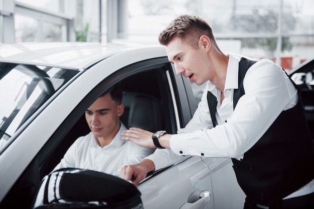 두 사람이 자동차에 대항하여 쇼룸에 서있다. 고객에게 자동차를 판매하는 소송에서 영업 관리자의 근접. 판매자는 고객에게 열쇠를 제공합니다.