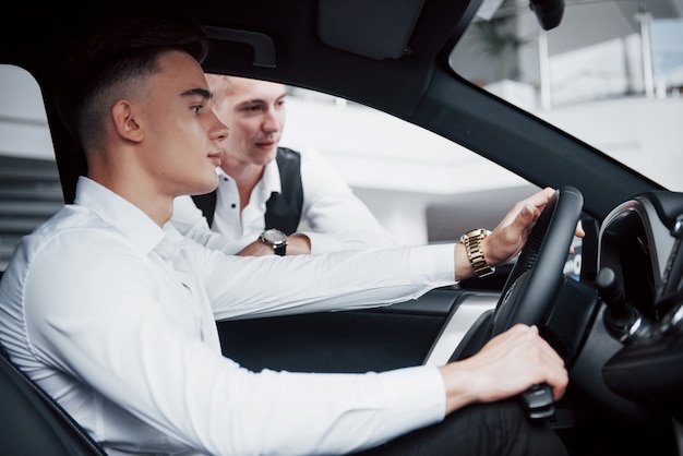 Two men stand in the showroom against cars. Close-up of a sales manager in a suit that sells a car to a customer. The seller gives the key to the customer.