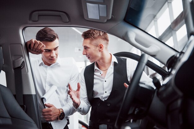 두 사람이 자동차에 대항하여 쇼룸에 서있다. 고객에게 자동차를 판매하는 소송에서 영업 관리자의 근접. 판매자는 고객에게 열쇠를 제공합니다.