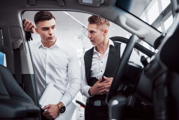 Two men stand in the showroom against cars. Close-up of a sales manager in a suit that sells a car to a customer. The seller gives the key to the customer.