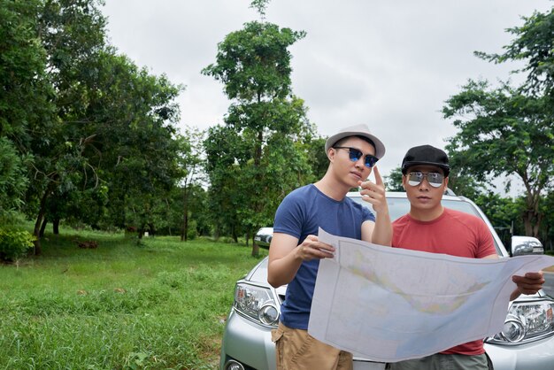 Two men in sunglasses standing by car, holding large map and pointing