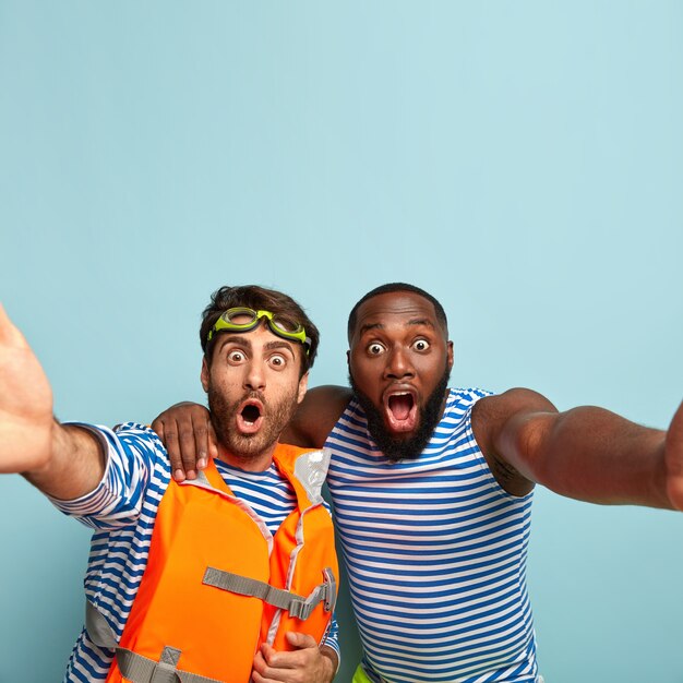 Free photo two mixed race men stare with shocked expressions, make selfie portrait, embrace, keep jaws dropped, spend leisure time at seaside