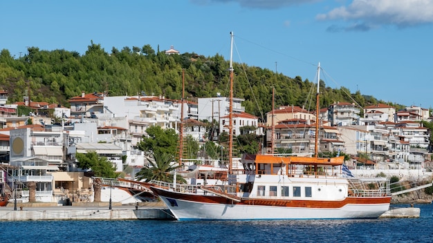 Two moored sailboats near a pier in Neos Marmaras, buildings located on a hill with multiple greenery, Greece