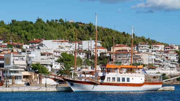 Two moored sailboats near a pier in Neos Marmaras, buildings located on a hill with multiple greenery, Greece