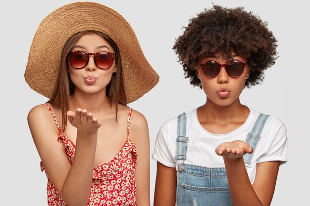 Two multiethnic sisters blow airkiss at camera, wears trendy shades, summer clothing