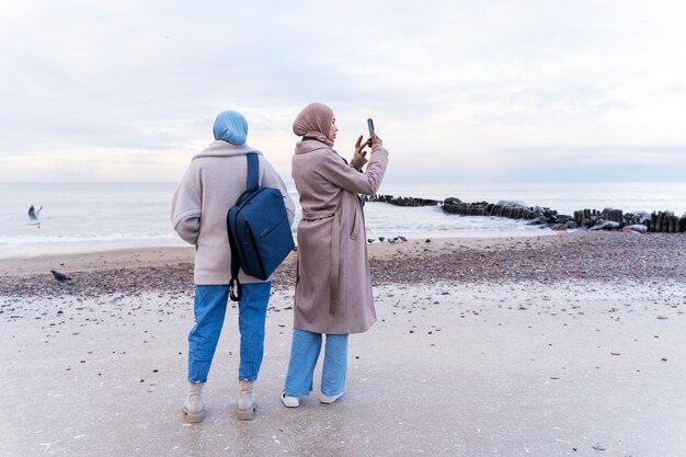 Two muslim women taking photos with smartphone on the beach while traveling