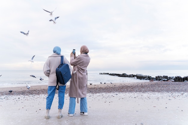 Two muslim women taking photos with smartphone on the beach while traveling