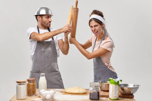 Two opponents at kitchen. Woman and man cooks struggle with kitchen utensils, compete who cooks better, make dough for baking pie, wear aprons, isolated over white wall. Culinary battle