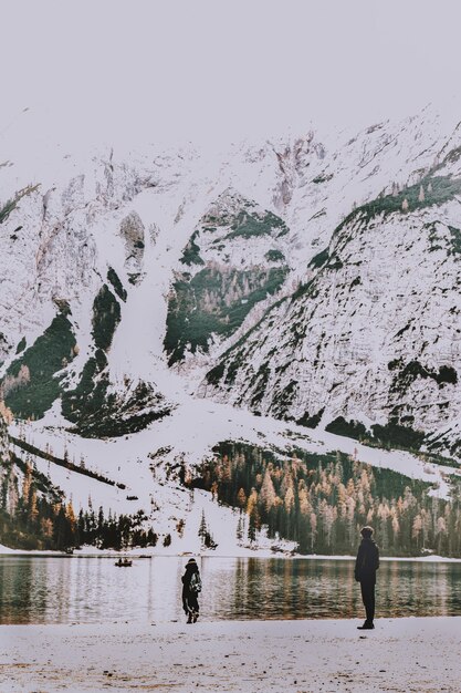 Two People Standing on Shore Overlooking Body of Water and Snow Covered Mountain