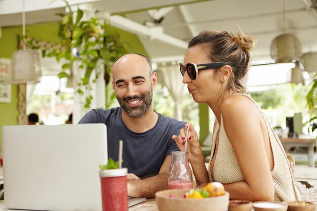 Two people watching video online or viewing pictures on Internet, using wi-fi on laptop computer during lunch. Happy bearded man and stylish woman in shades relaxing at cafe with notebook.