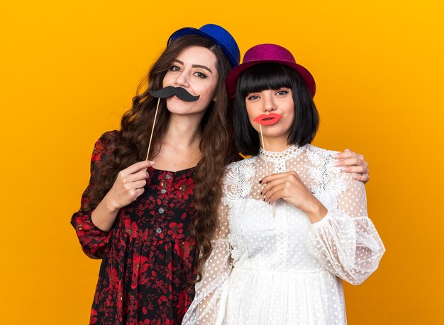 Two pleased young party women wearing party hat looking at front, holding fake mustache and lips