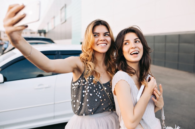 Two pretty fashionable girls making selfie-portrait on parking.