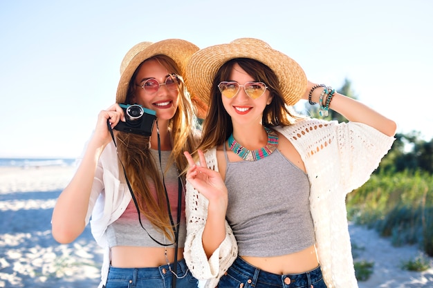 Two pretty funny sister girls making selfie on vintage camera, posing on the beach, party and vacation mood, crazy positive feeling, summer bright clothes sunglasses and hats.