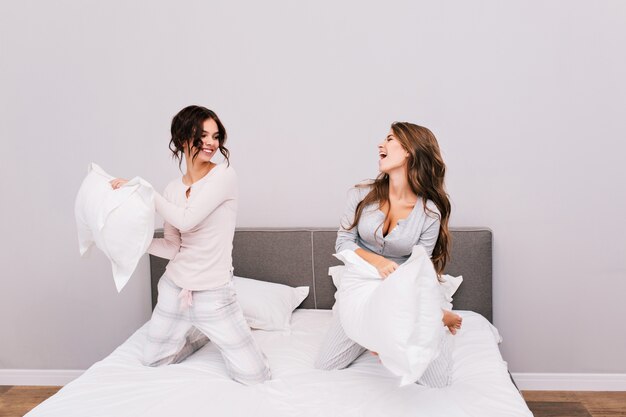 Две красивые девушки в пижамах дрались подушками на кровати.