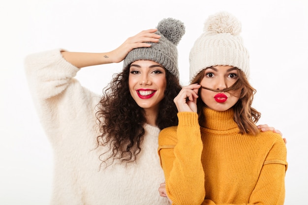 Two pretty girls in sweaters and hats posing together  over white wall