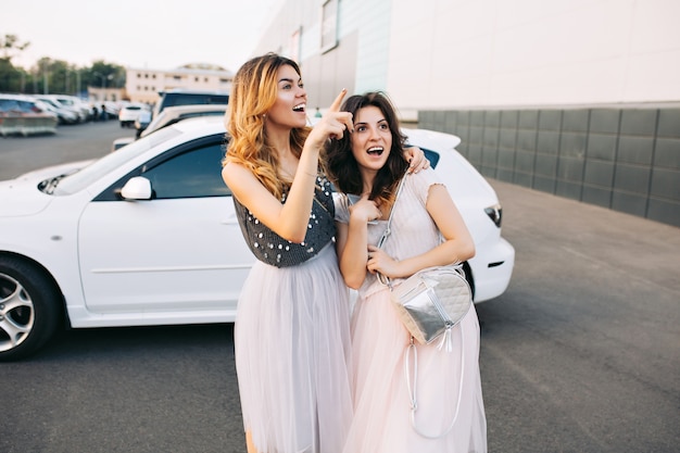 Two pretty girls in  tulle skirts having fun on parking. They looking surprised and excited far away.