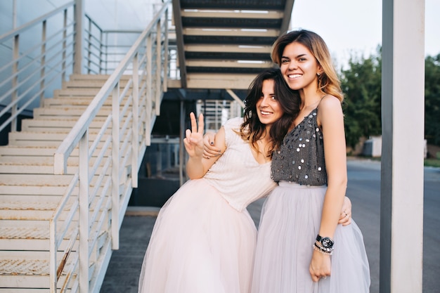 Two pretty girls in tulle skirts outside near grey stairs. They hugging and smiling .