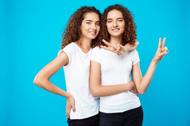 Two pretty girls twins smiling, showing peace over blue wall