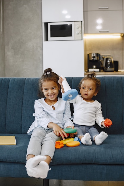 Двое симпатичных малышей сидят на диване и играют с игрушками. Афро-американские сестры играют дома.
