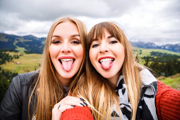 Two pretty traveler girls making funny selfie at mountains. Showing long tongues, going crazy.