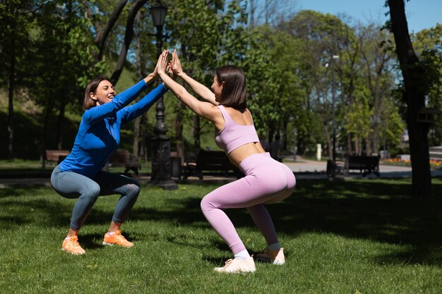 Two pretty woman in sport wear on grass in park at sunny day doing workout squats support each other happy emotions