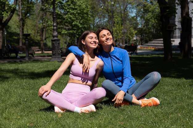 Две красивые женщины в спортивной одежде на траве в парке в солнечный день, занимаясь йогой, обнимают друг друга с улыбкой на лице