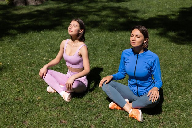 Two pretty woman in sport wear on grass in park at sunny day doing yoga poses, catch sun rays