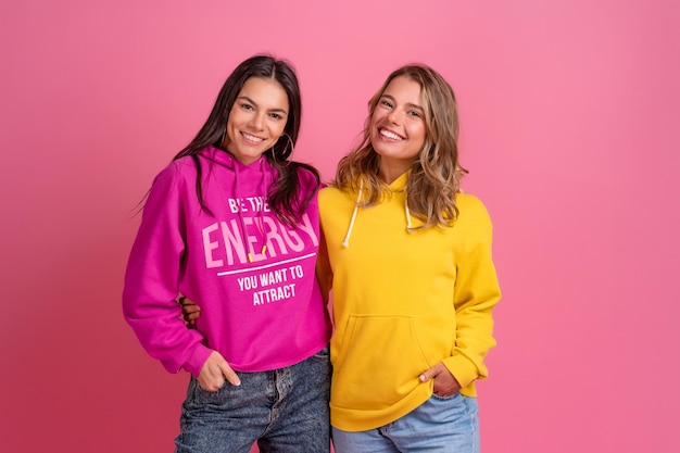 Two pretty women friends embracing having fun together blond and brunette wearing hoodie posing on pink background isolated