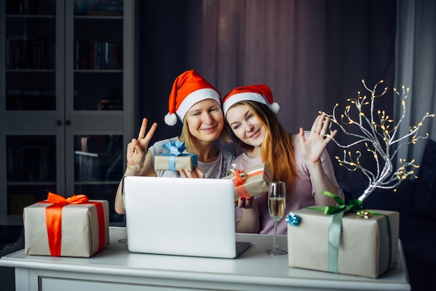 산타클로스 모자를 쓴 예쁜 여성 두 명이 화환과 선물 사이에 노트북을 들고 테이블에 앉아 메리 크리스마스를 기원합니다. 2022년 새해 전야에 인터넷을 통해 친구 및 가족과 통신합니다.