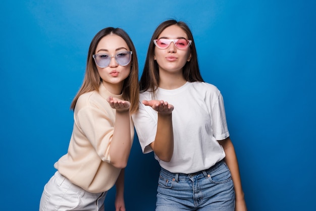 Две симпатичные молодые девушки, одетые в летнюю одежду, стоят и отправляют поцелуй, изолированные на синей стене